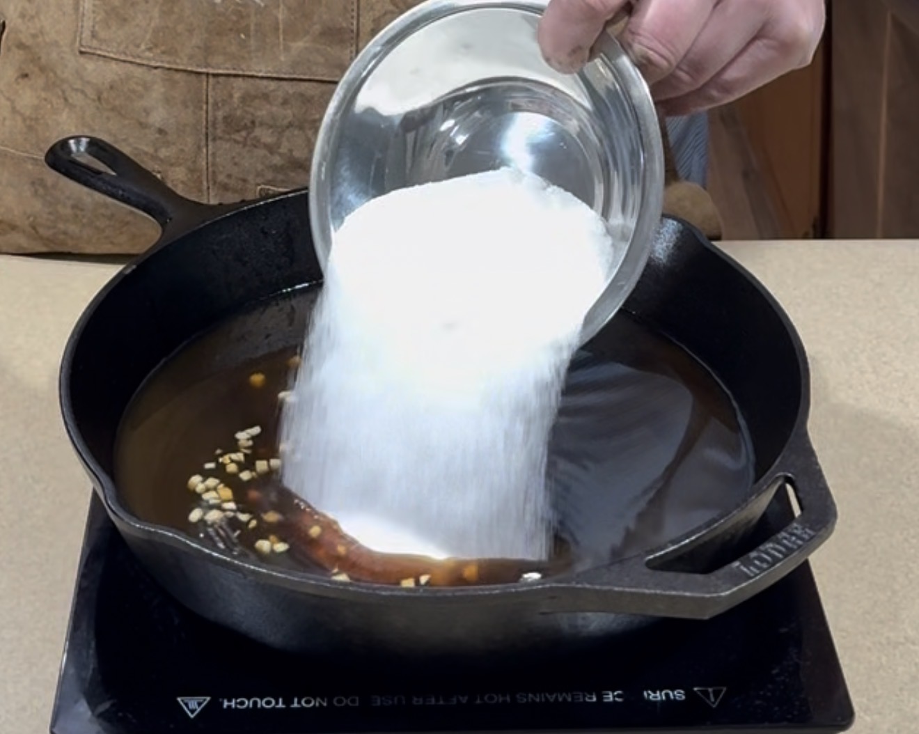 A hand pours a bowl of white granulated sugar into a cast iron skillet containing a dark liquid with chopped garlic pieces on an electric stovetop.
