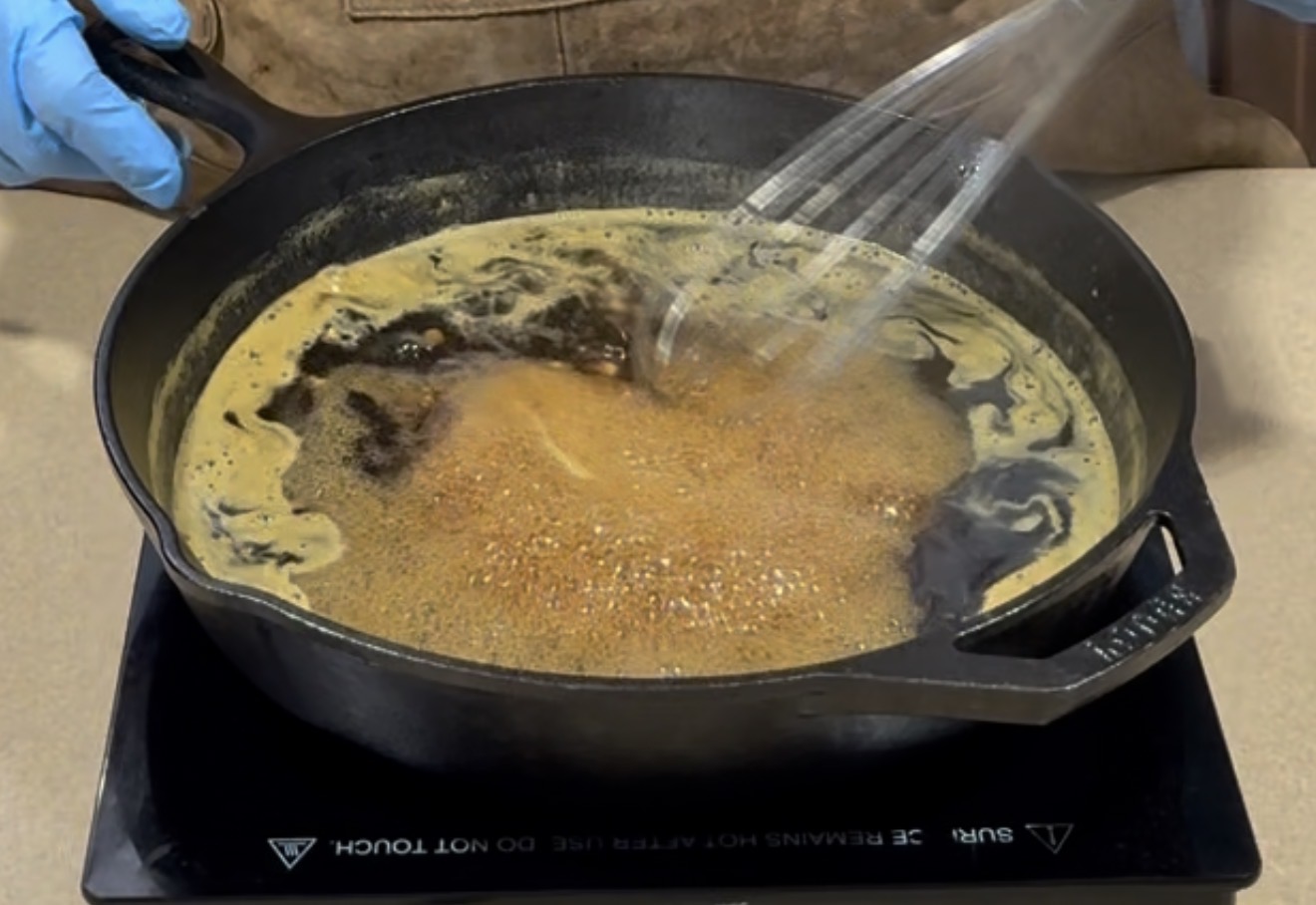 A person wearing a blue glove on one hand uses a whisk to stir a frothy, dark liquid in a black cast iron pan set on an induction cooktop.