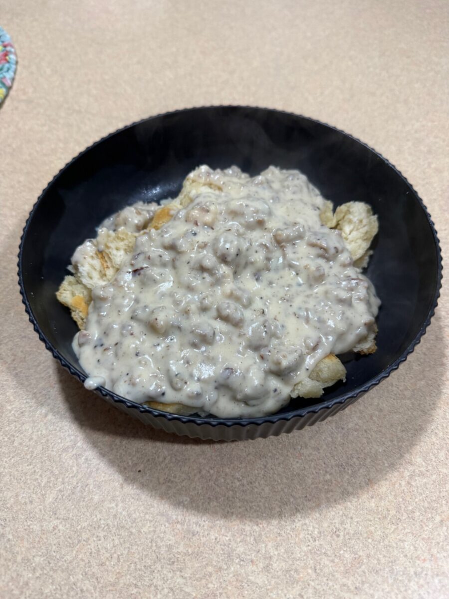 A black bowl filled with biscuits topped with creamy sausage gravy sits on a light-colored countertop. Steam rises from the dish, indicating it is freshly made and hot.