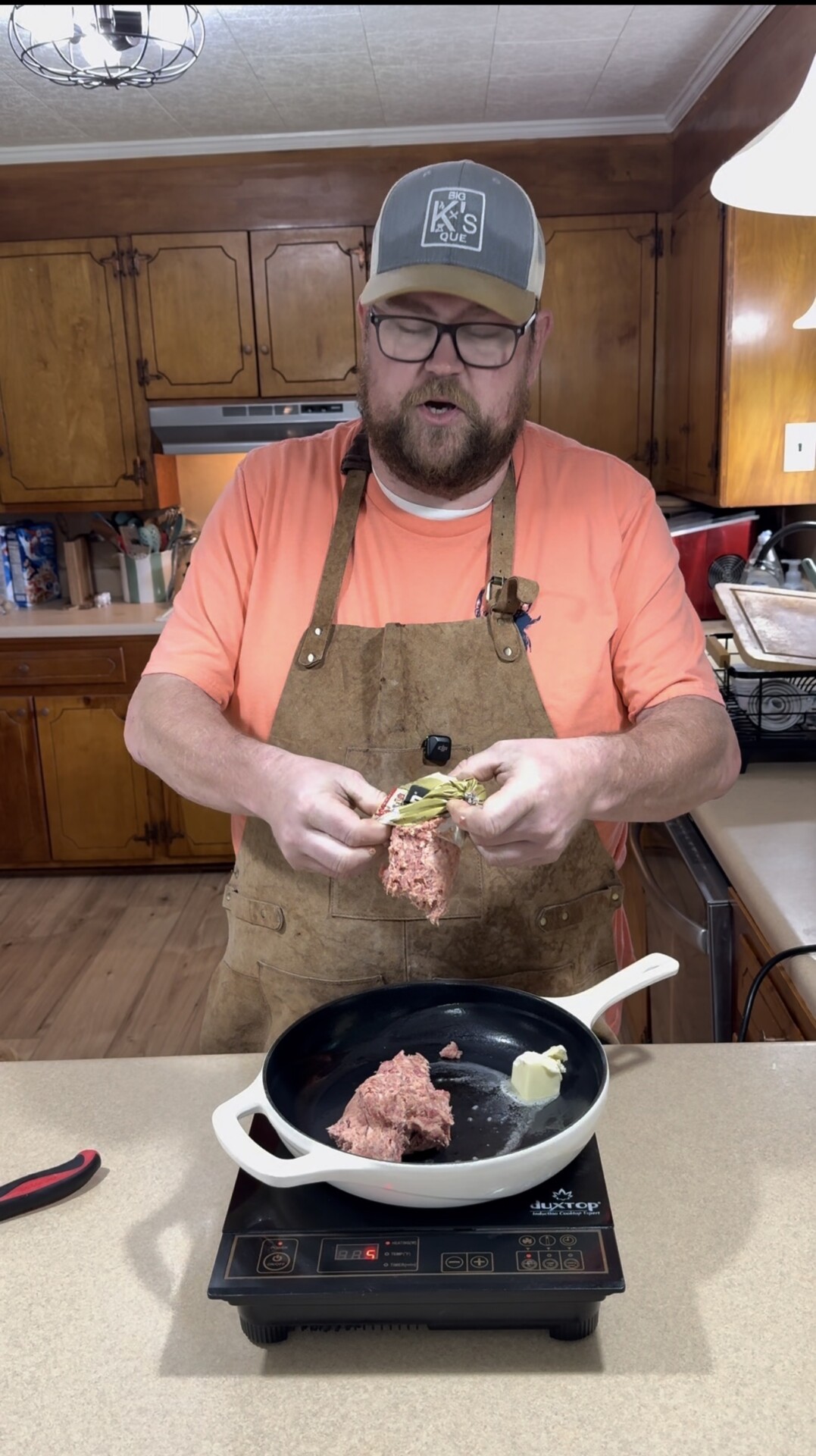 A man wearing a cap, glasses, and apron stands in a kitchen, adding ground meat and butter to a pan on a stovetop. Wooden cabinets and kitchen items are visible in the background.