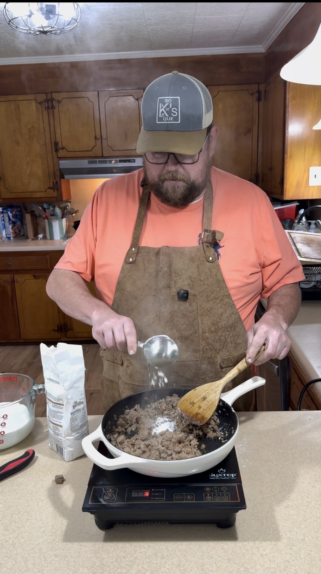 A man wearing a tan apron and baseball cap is cooking ground meat in a white skillet on a stovetop, sprinkling flour into the pan. Measuring cups, flour, and utensils are visible on the kitchen counter.