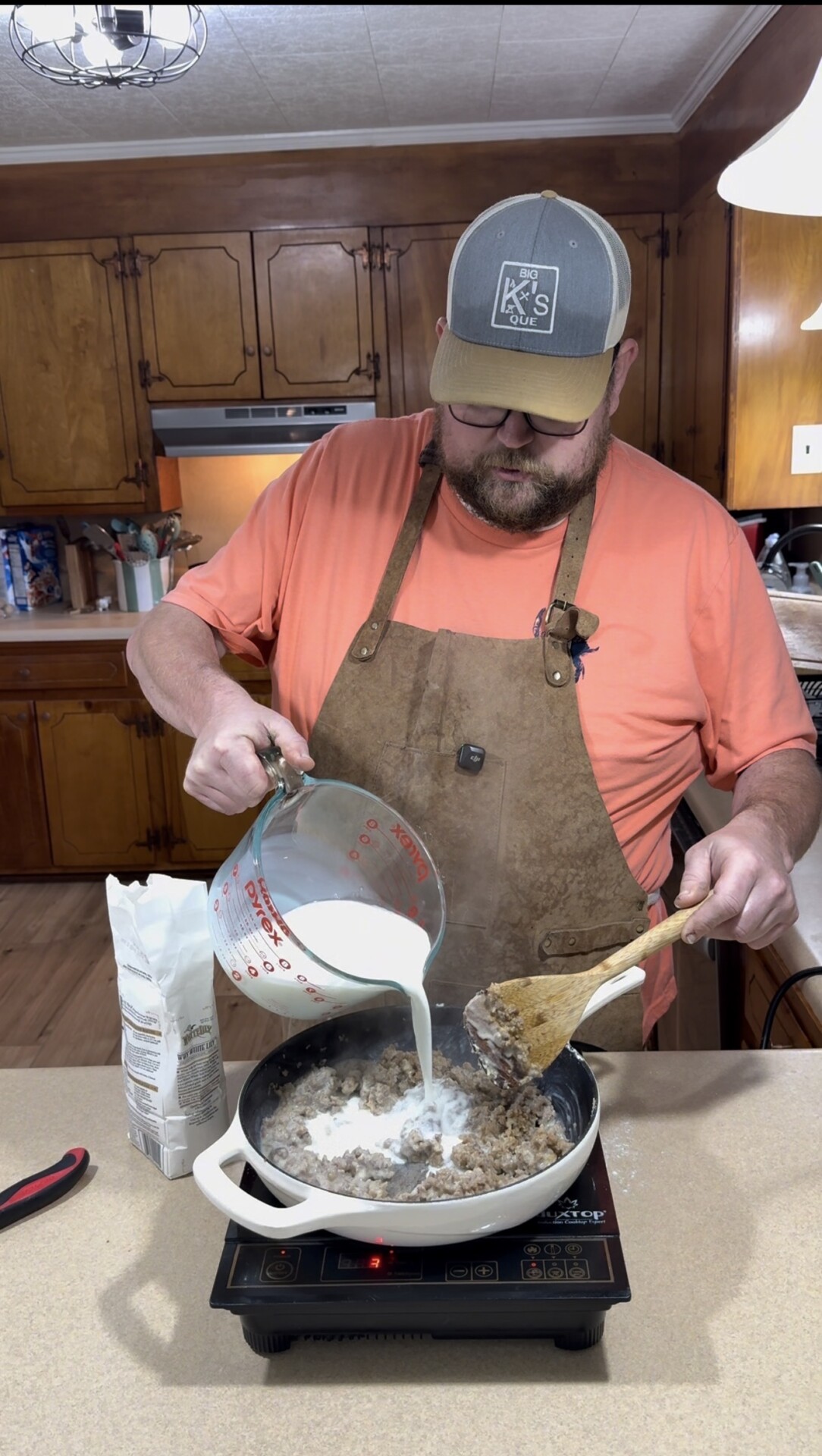 A man in an apron and a baseball cap pours milk from a measuring cup into a skillet of ground meat on a stovetop, stirring with a wooden spoon in a kitchen with wood cabinets.