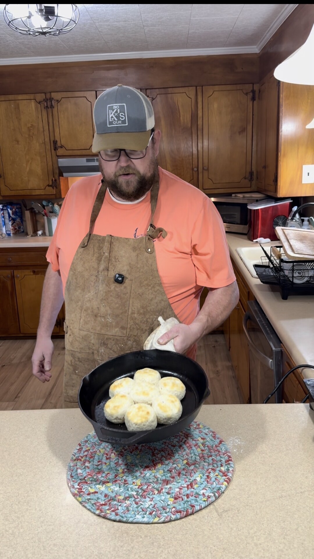 A man wearing a brown apron and a cap holds a cast iron skillet with freshly baked biscuits in a kitchen, placing it on a colorful trivet on the countertop.