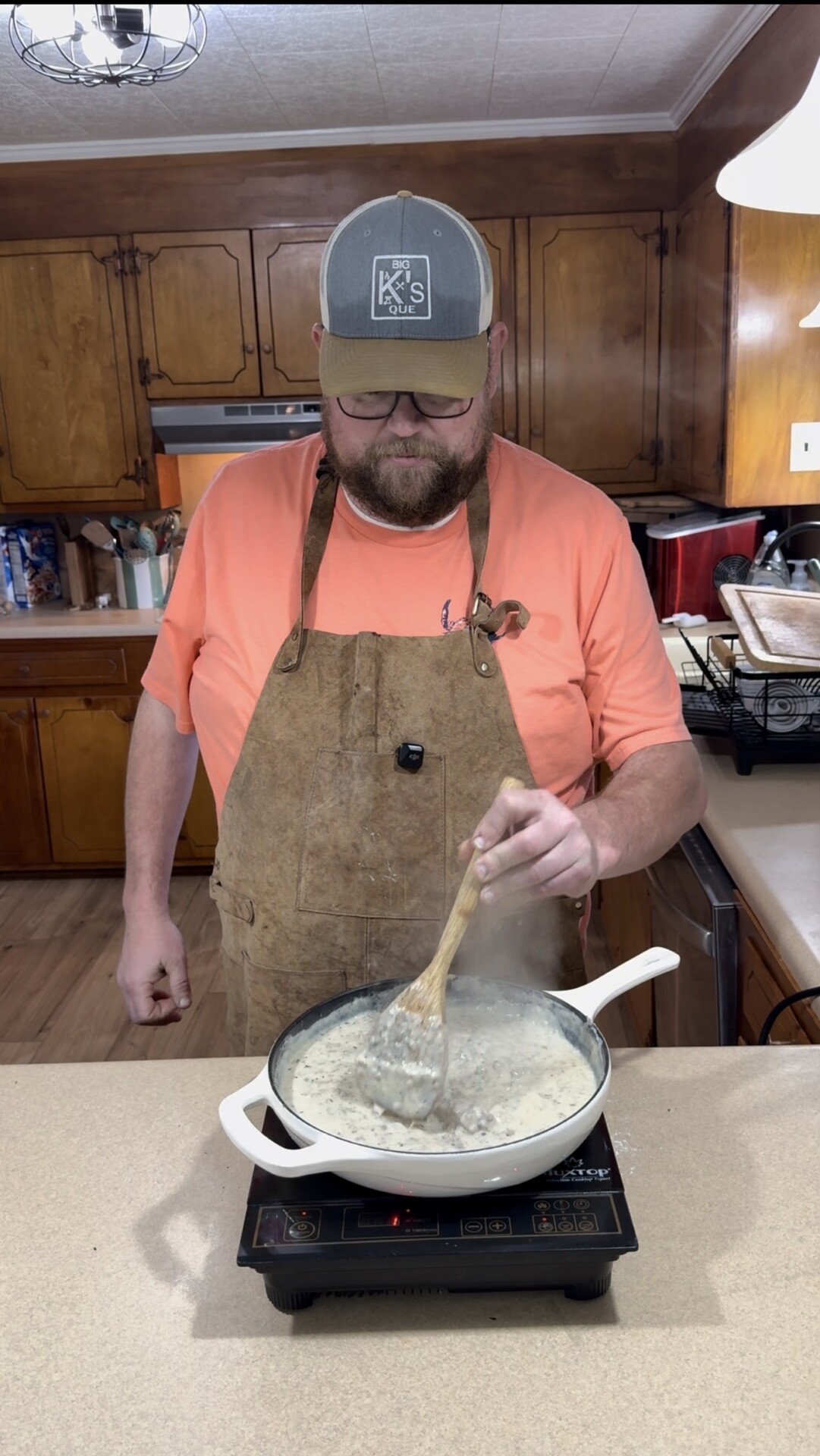 A man wearing a brown apron, orange shirt, and baseball cap stirs a creamy mixture in a white pan on an electric cooktop in a kitchen with wooden cabinets. Steam rises from the pan.