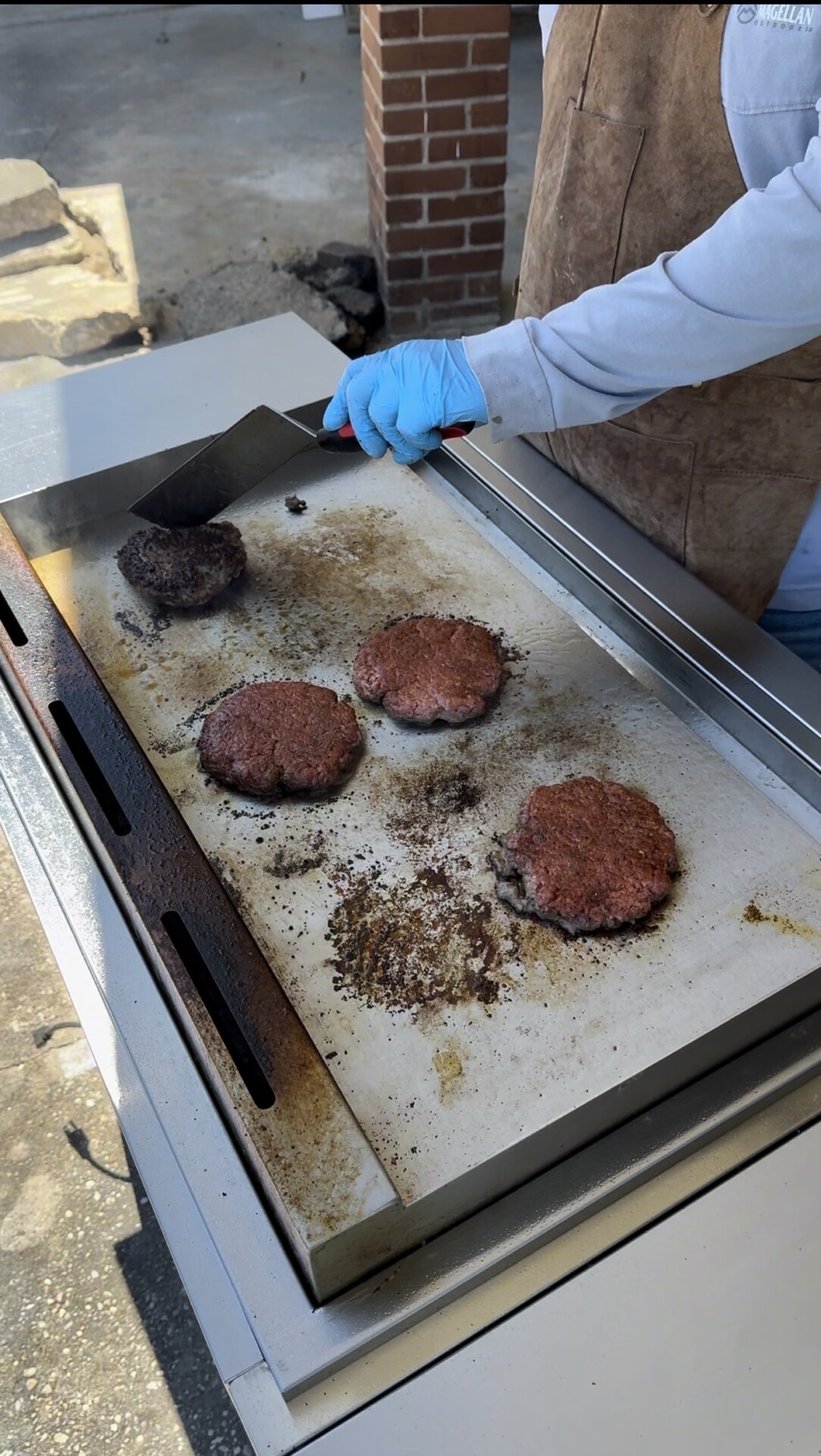 A person wearing a blue glove and apron uses a spatula to cook four hamburger patties on a flat-top outdoor grill; one patty is well-cooked while the others are still raw or partially cooked.
