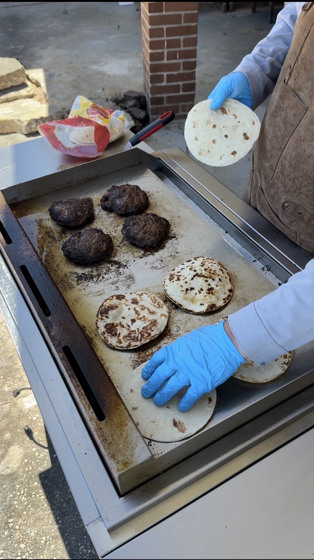 A person wearing blue gloves and a brown apron grills burger patties and toasts flour tortillas on an outdoor flat-top griddle. A bag of tortillas and a spatula rest nearby.