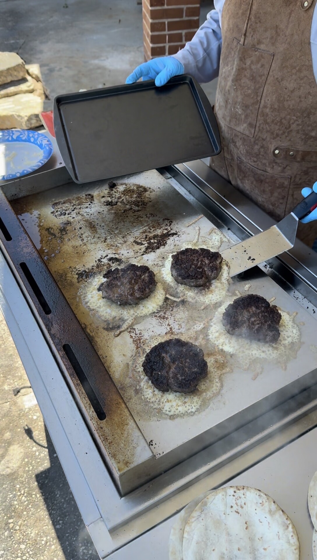 A person wearing blue gloves and a brown apron is cooking burger patties on a flat-top grill outdoors, using a spatula to flip the patties. Four patties are visible, sizzling and slightly charred.