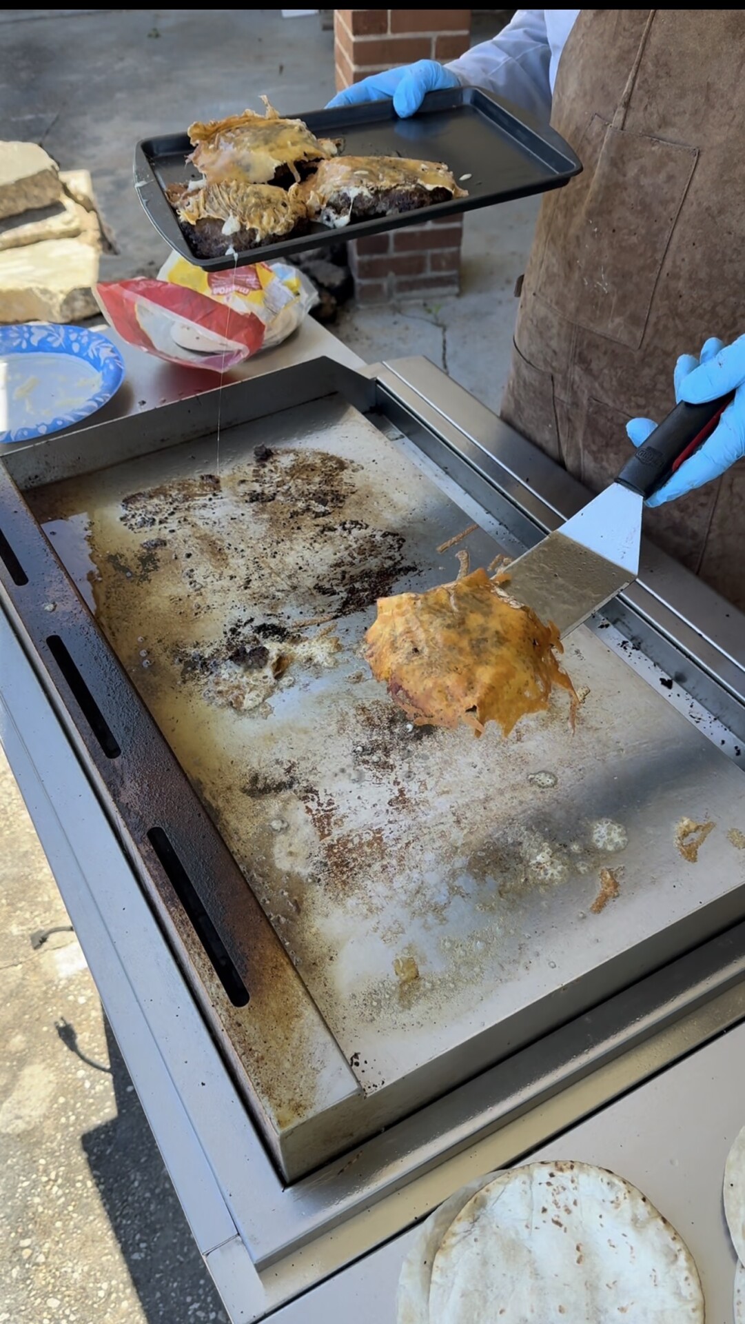 A person wearing blue gloves uses a spatula to lift a cooked, cheesy patty from a griddle, with another patty on a tray nearby. Tortillas and plates are visible on the side.