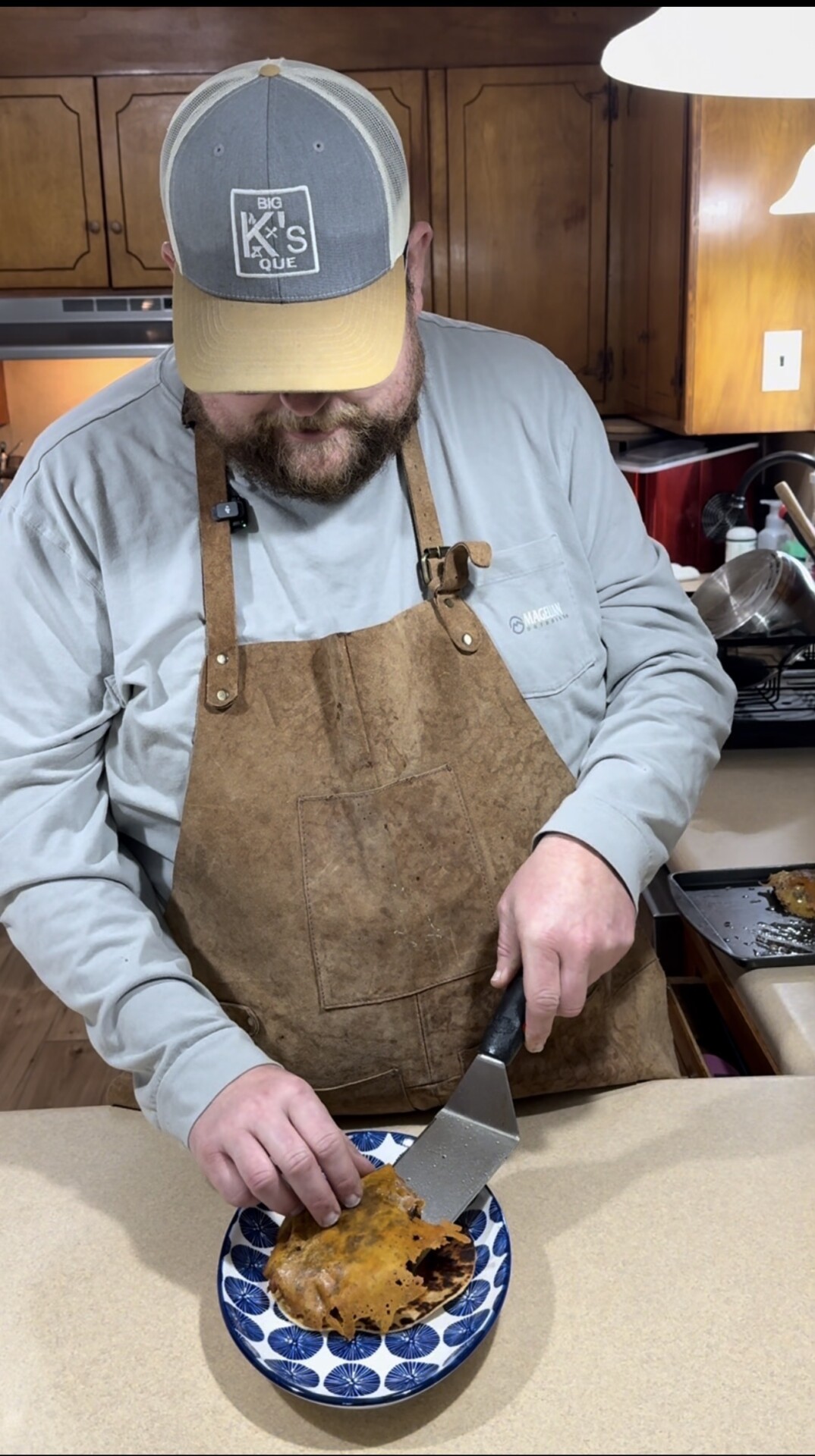 A man wearing a brown apron and a gray cap is slicing a cooked piece of food on a blue and white plate in a kitchen. He is focused on his task, standing by a countertop with a tray in the background.