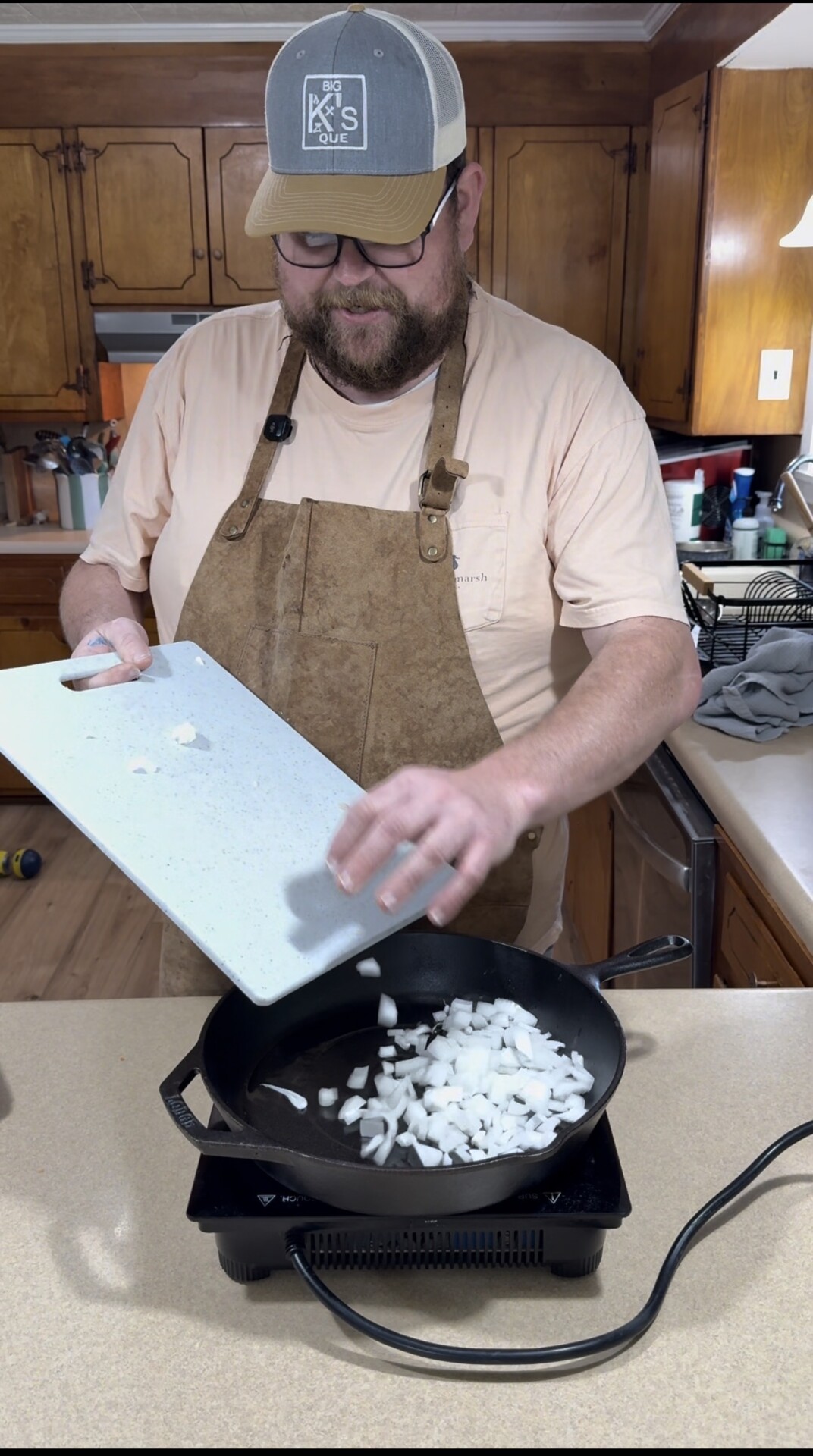A man wearing a brown apron and cap pours chopped onions from a cutting board into a cast iron skillet on a kitchen counter. Wooden cabinets and kitchen utensils are visible in the background.