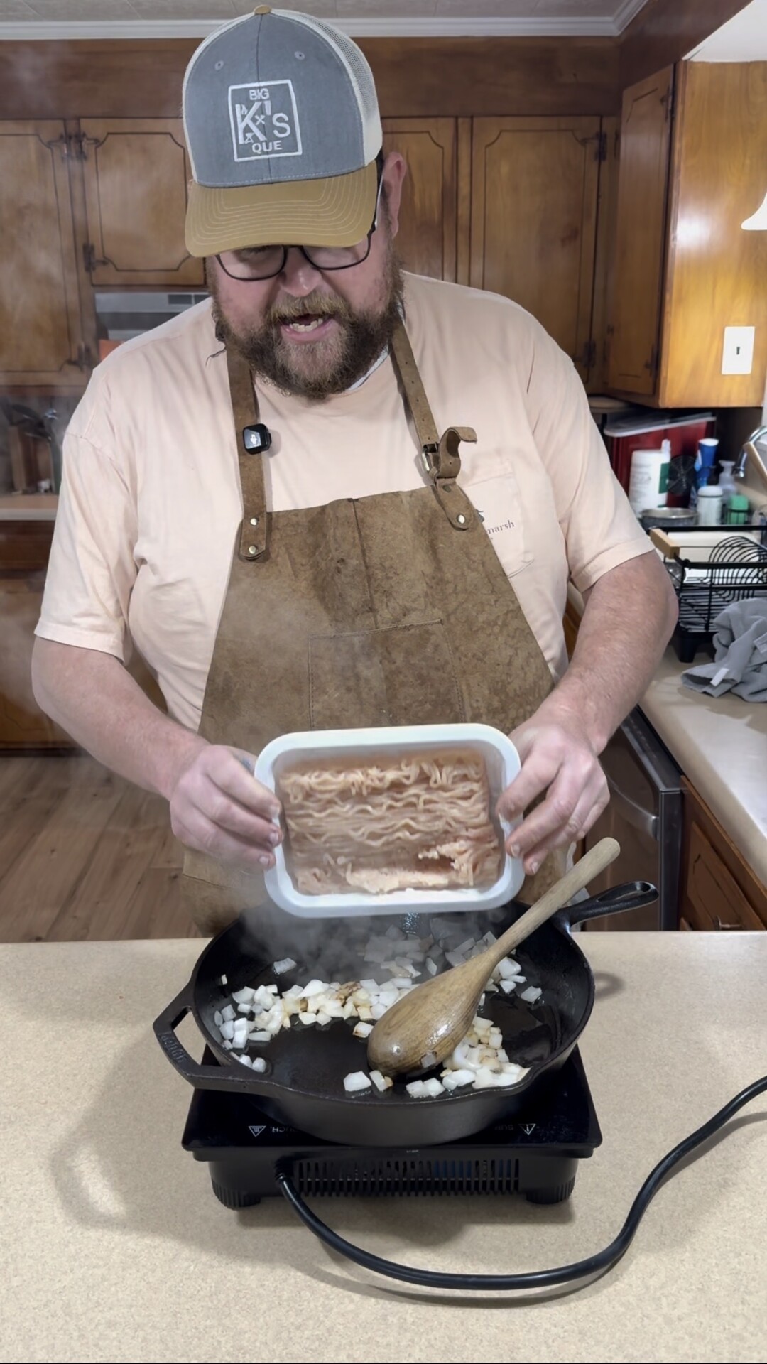 A man wearing a cap, glasses, apron, and light shirt is cooking in a kitchen. He holds a container of raw ground meat over a pan with onions on a stovetop, preparing to add the meat. A wooden spoon rests in the pan.