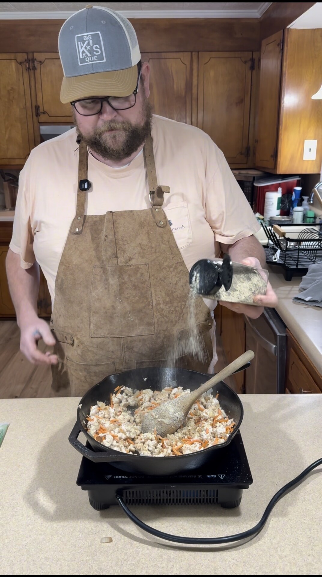 A man wearing a brown apron and a cap pours seasoning from a container into a skillet with chopped vegetables and meat on a stovetop in a home kitchen.