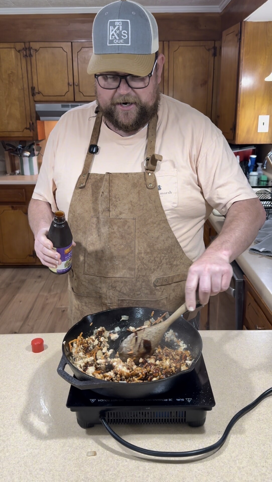 A man in an apron cooks at a stove in a kitchen, stirring food in a skillet with a wooden spoon while holding a bottle of sauce. The kitchen has wooden cabinets and various utensils on the counter.