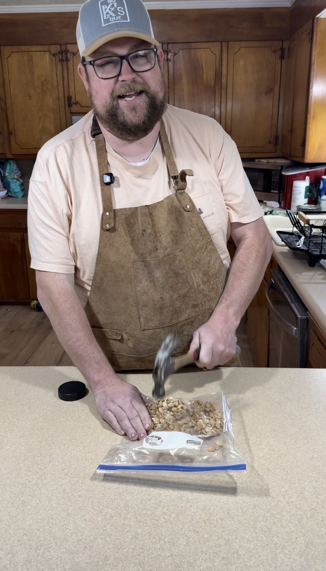 A man wearing an apron and a cap is smiling while using a meat tenderizer to crush nuts inside a plastic bag on a kitchen counter. Wooden cabinets and kitchen items are visible in the background.