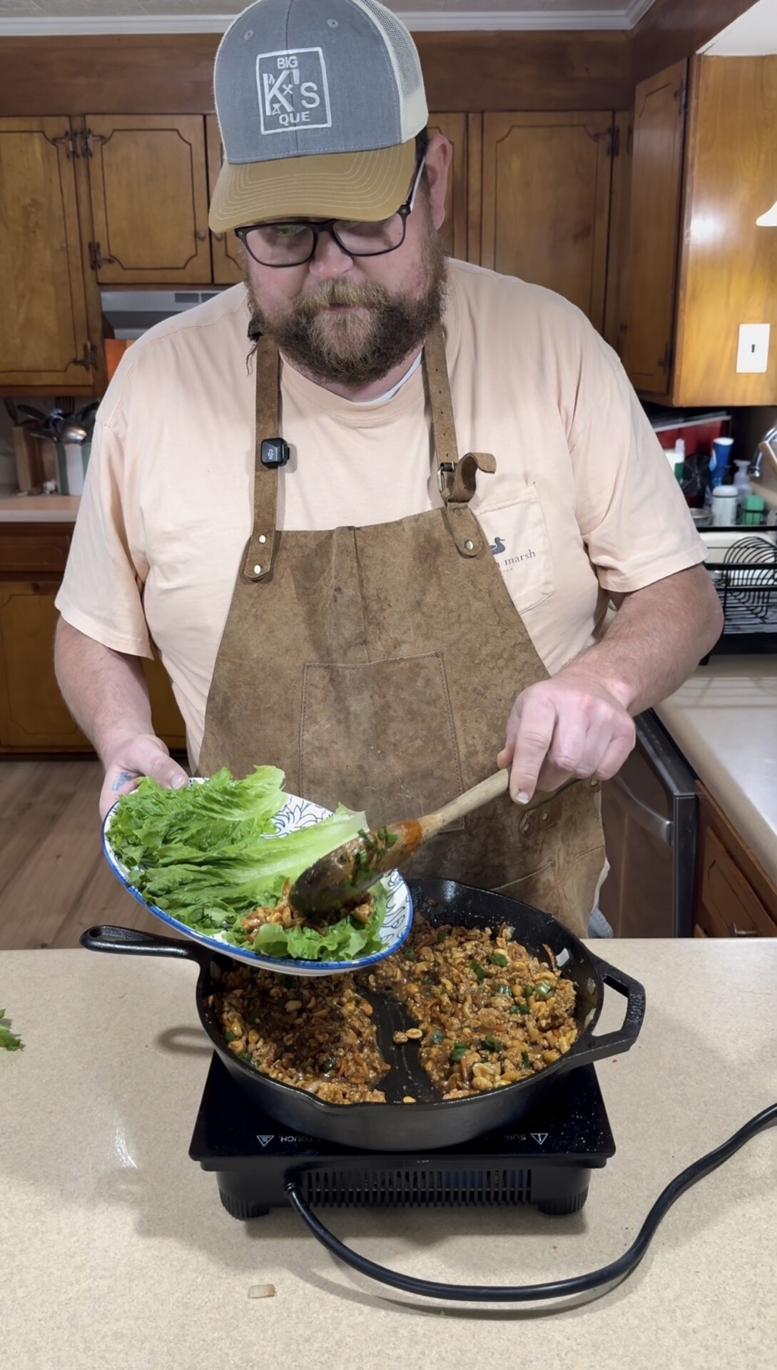 A man wearing a hat, glasses, and a brown apron is spooning a cooked mixture from a skillet into a bowl of lettuce leaves in a kitchen.