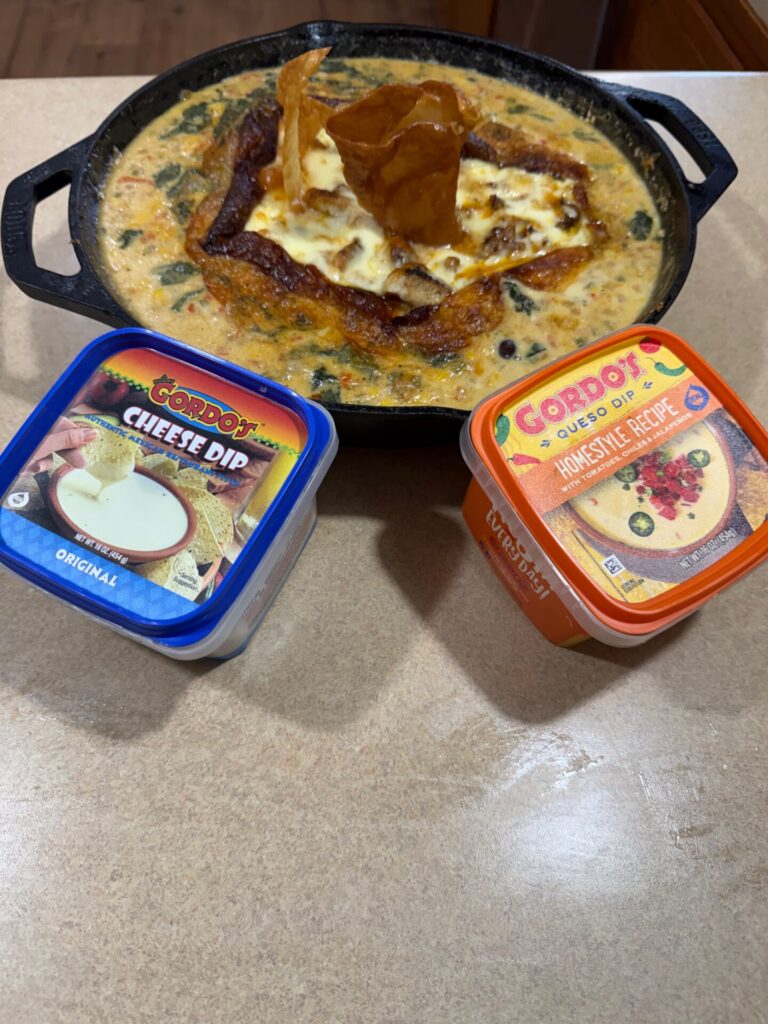 A skillet of cheesy baked dip with a crispy topping is on a counter, next to containers of Qordos Cheese Dip and Qordos Queso Dip Homestyle Recipe.