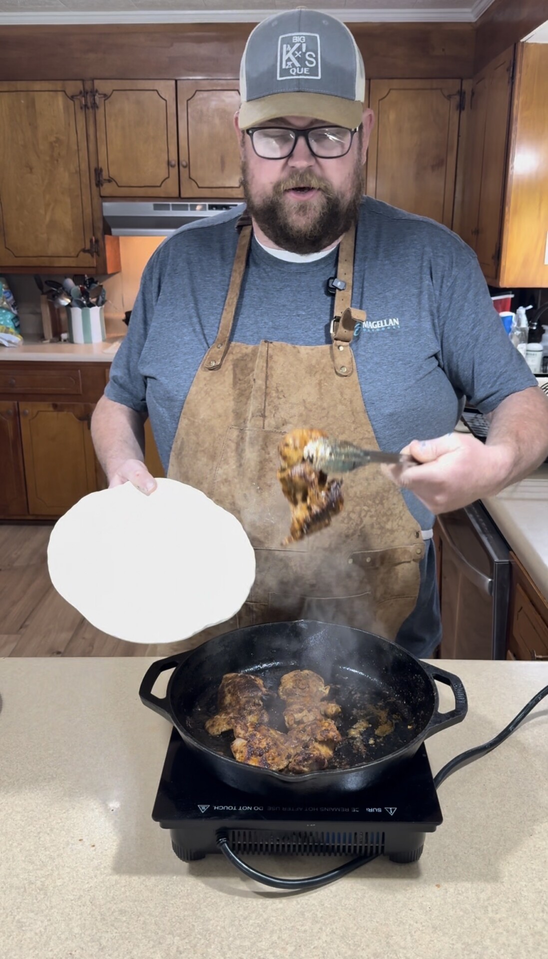 A man wearing glasses, a cap, and an apron stands in a kitchen, holding a plate and serving food from a steaming pan on a stovetop. The kitchen features wooden cabinets and countertops.