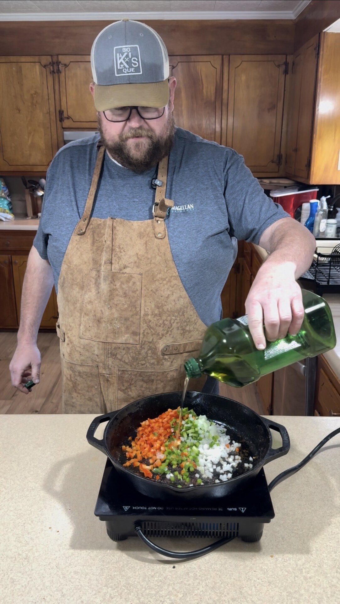 A bearded man wearing glasses, a tan apron, and a cap pours olive oil from a bottle into a skillet with chopped onions, celery, and carrots on a stovetop in a home kitchen.