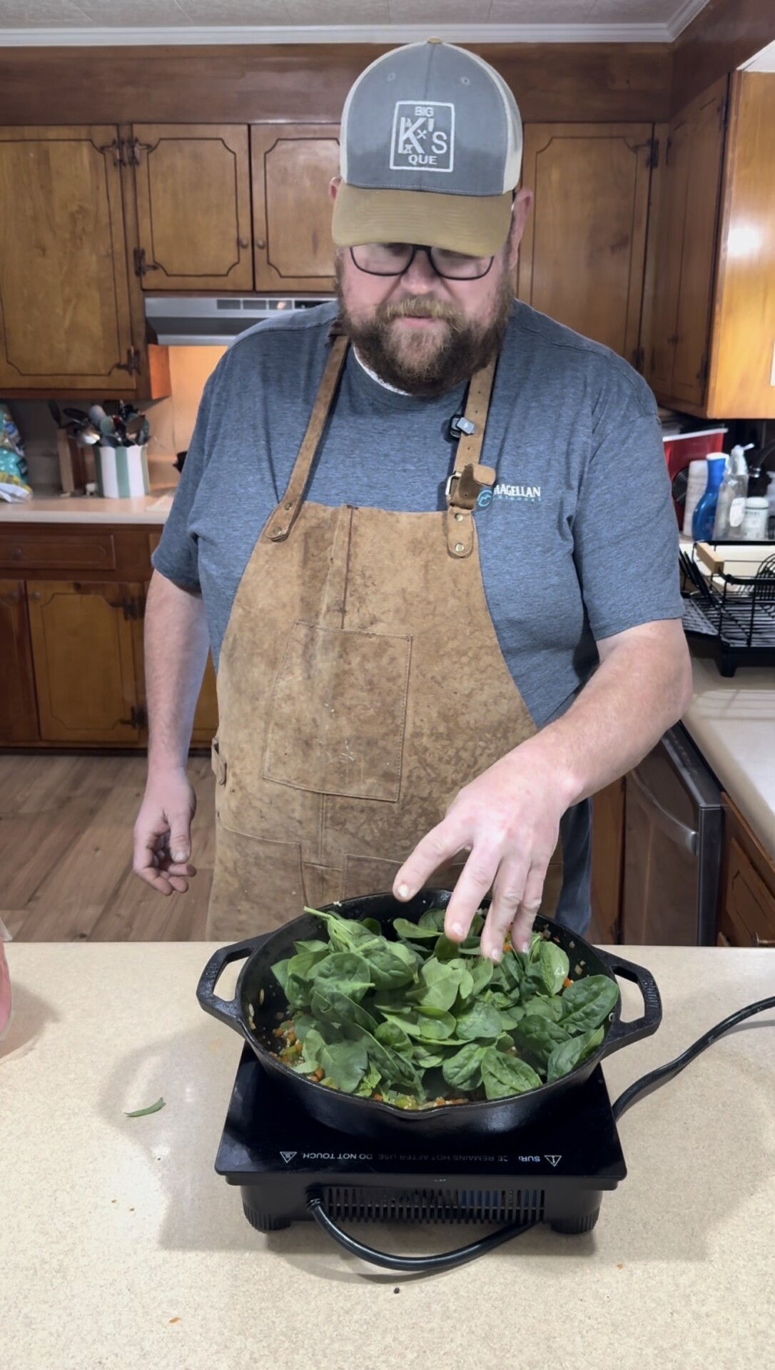 A man wearing a brown apron, gray t-shirt, and cap is cooking in a kitchen, placing fresh spinach into a skillet on an electric stove. Wooden cabinets and a countertop with kitchen items are visible in the background.