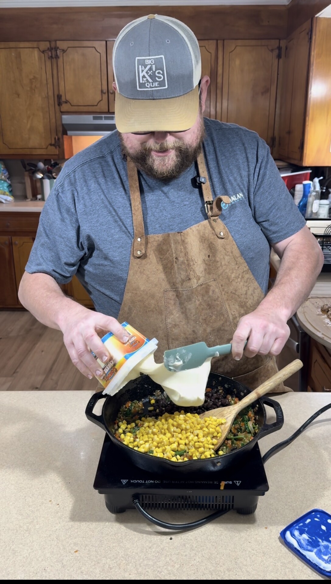 A man in a gray shirt, brown apron, and cap is smiling while adding a container of mayonnaise to a skillet filled with corn, black beans, and vegetables on a stove in a kitchen.