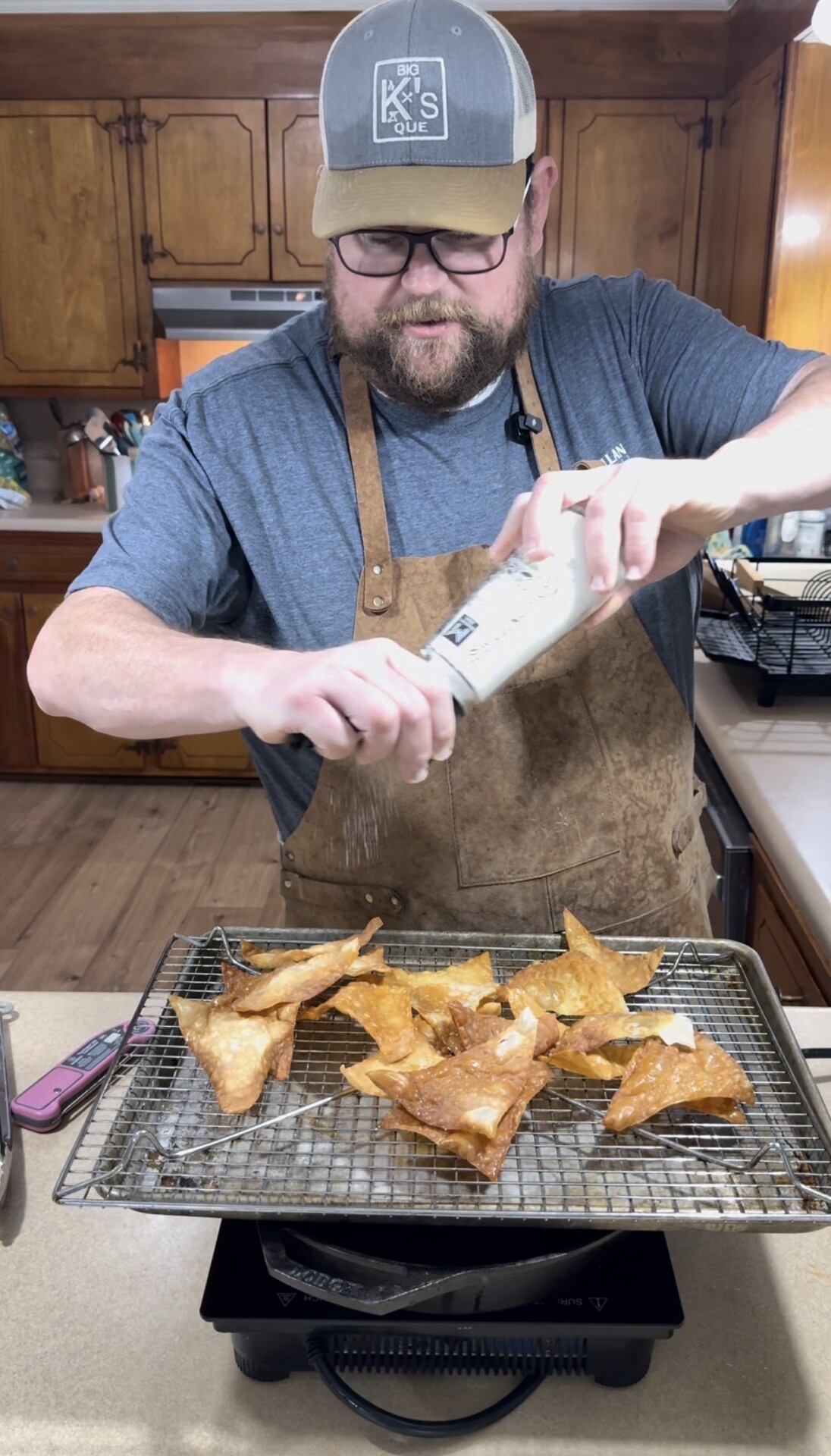A man in a blue shirt and brown apron sprinkles seasoning on fried tortilla chips on a wire rack in a kitchen with wooden cabinets.