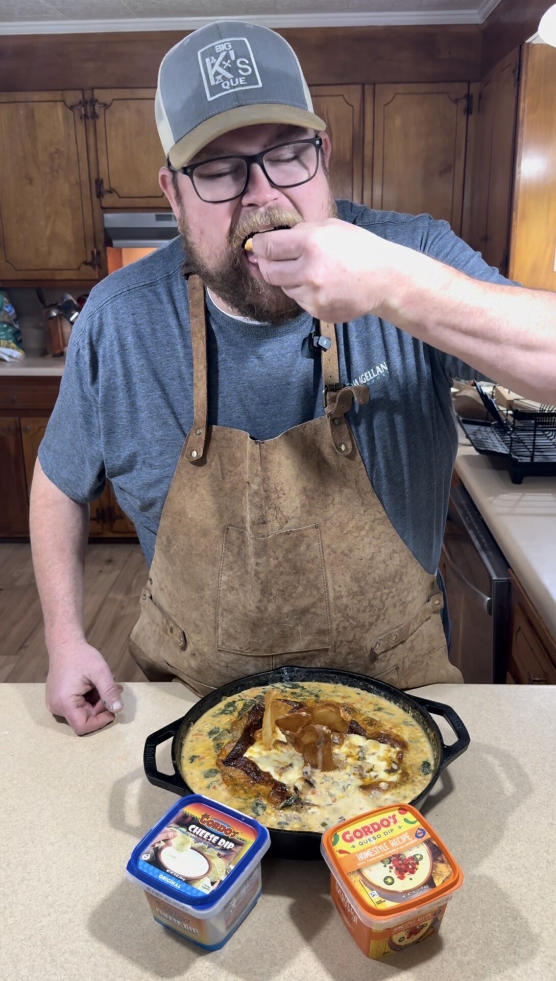 A bearded man in a cap, glasses, and a brown apron is standing in a kitchen, eating a cheesy dip from a skillet on the counter. Two containers of Cabot cheese products are in front of the skillet.