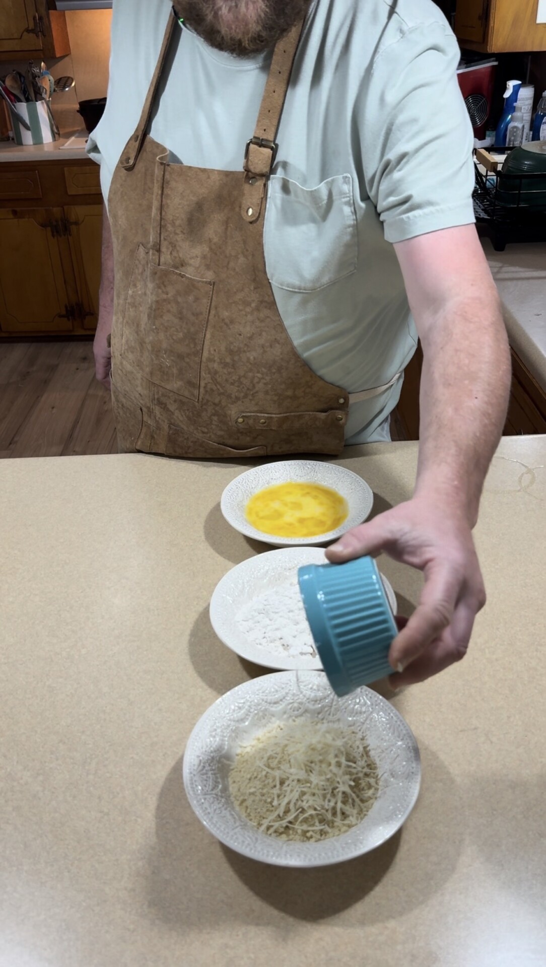 A person wearing a tan apron stands at a kitchen counter, holding a small blue bowl and sprinkling grated cheese onto a plate. Two other plates, one with flour and one with beaten eggs, are arranged in a row on the counter.