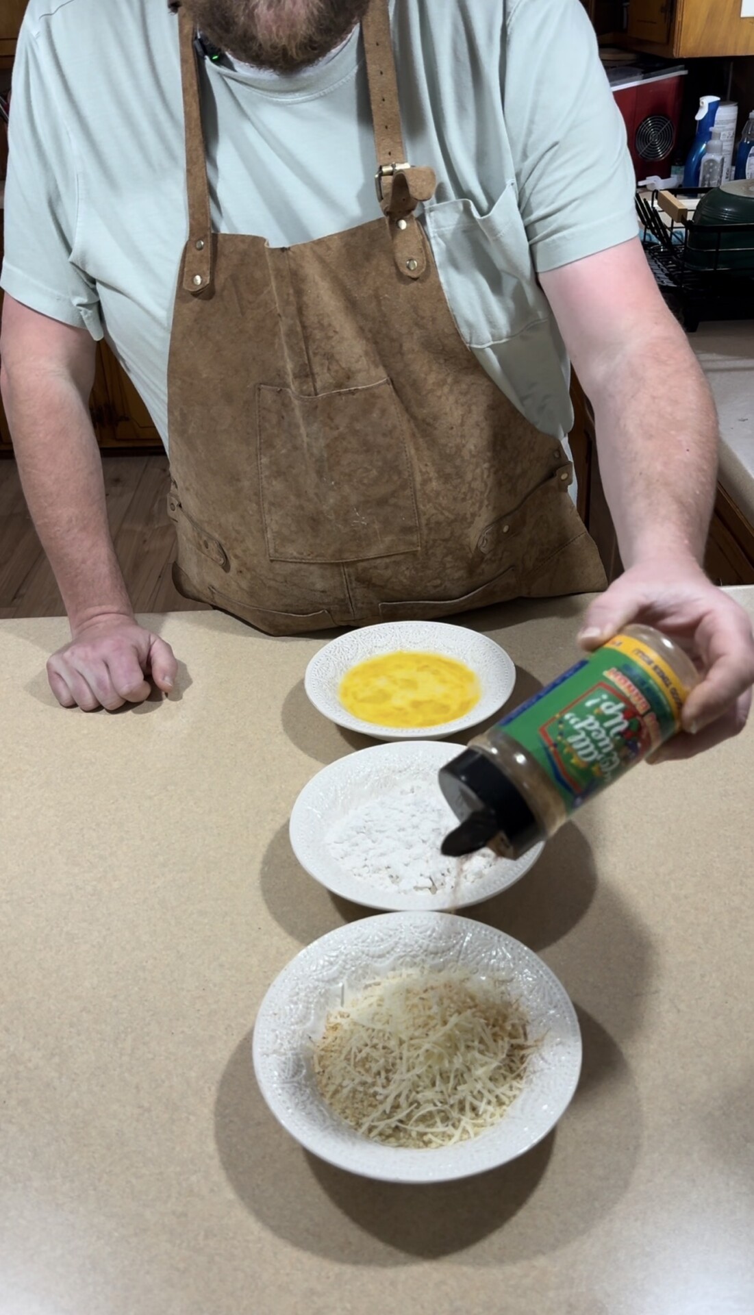 A person wearing a tan apron stands at a kitchen counter, holding a seasoning shaker over a plate of shredded cheese. Plates with egg wash and flour are also on the counter.