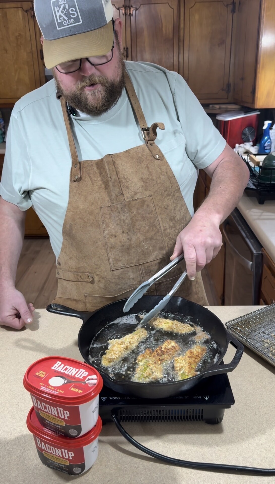 A man wearing an apron and hat uses tongs to fry breaded food in a cast-iron skillet on the stove. Two red containers labeled Bacon Up are on the counter beside him in a kitchen setting.