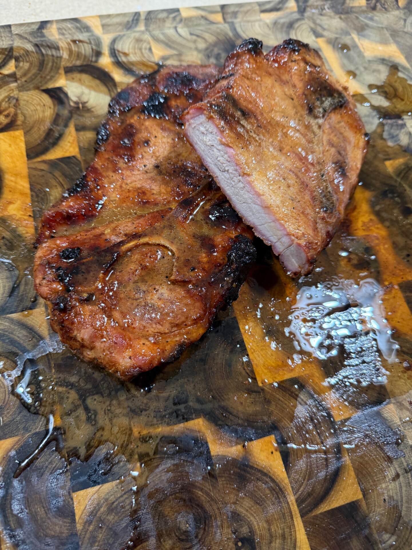 Grilled, juicy pork steak with a smoky char is sliced on a wooden cutting board, with visible meat juices pooling nearby.
