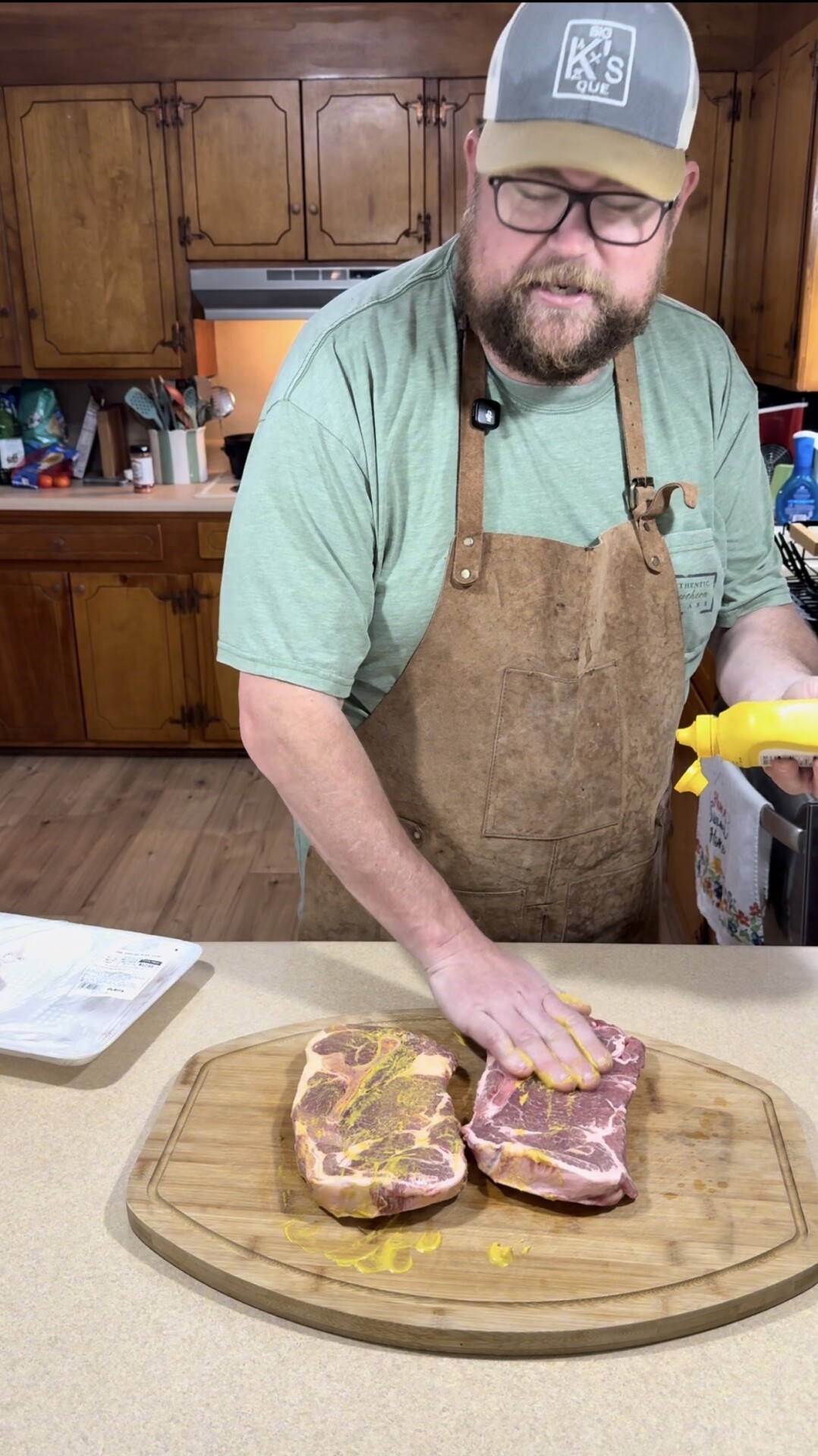 A bearded man wearing glasses, a cap, and a brown apron spreads yellow mustard onto two raw pieces of meat on a wooden cutting board in a kitchen.