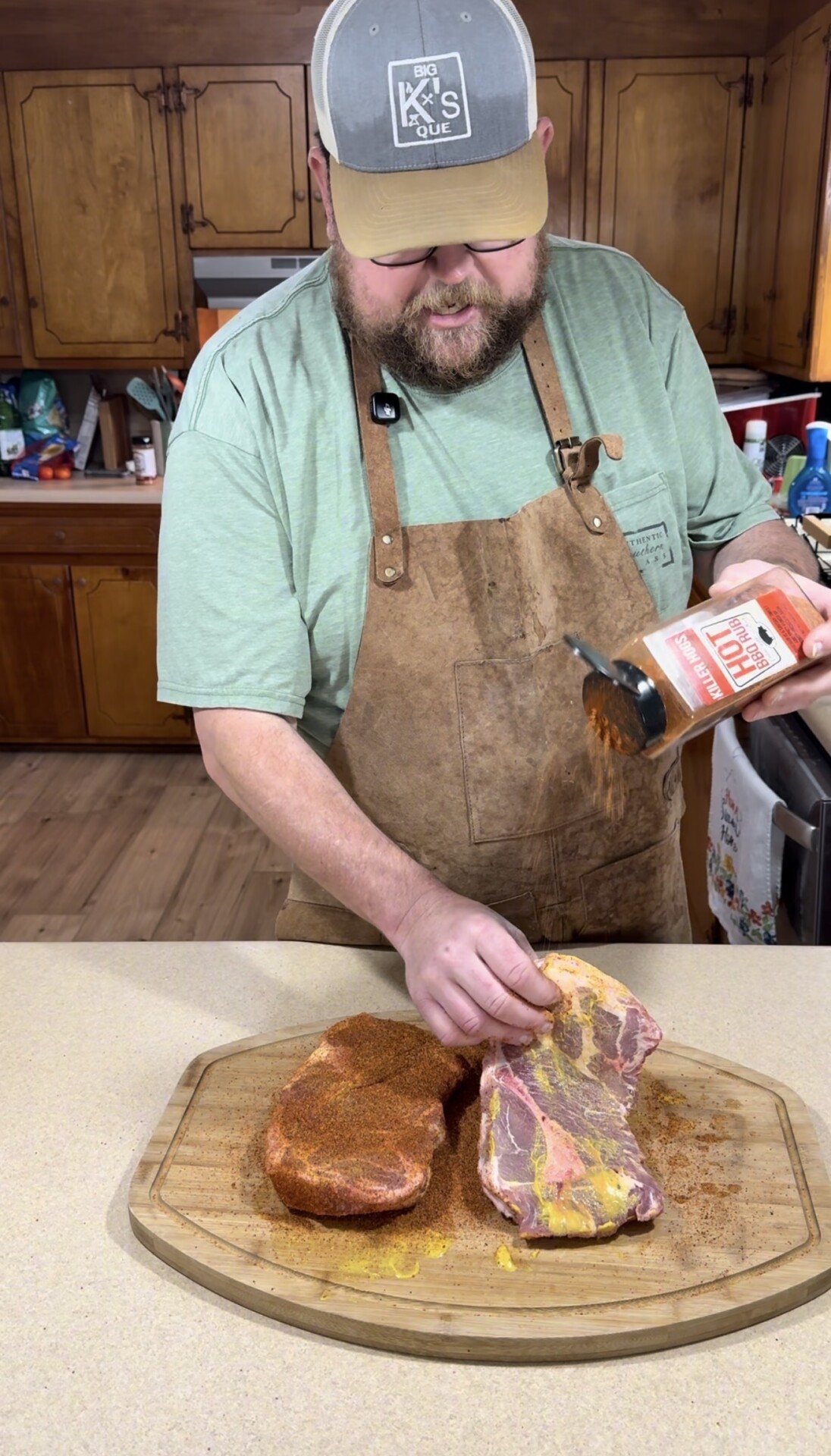 A man wearing a brown apron and a cap sprinkles seasoning onto two raw steaks on a wooden cutting board in a kitchen.