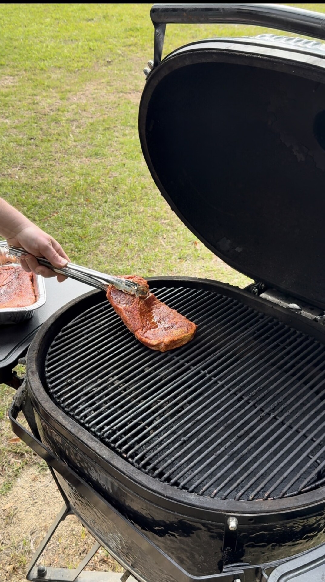 A person places a seasoned piece of meat onto the grill grate of a large black barbecue smoker outdoors, with green grass visible in the background.