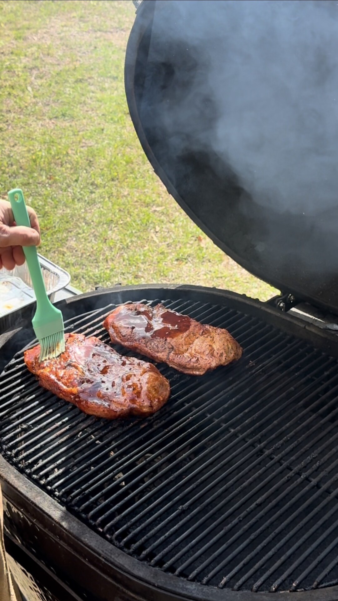 A person brushes sauce onto two large pieces of meat cooking on a grill, with smoke rising and the grill lid open. Grass and a tray are visible in the background.