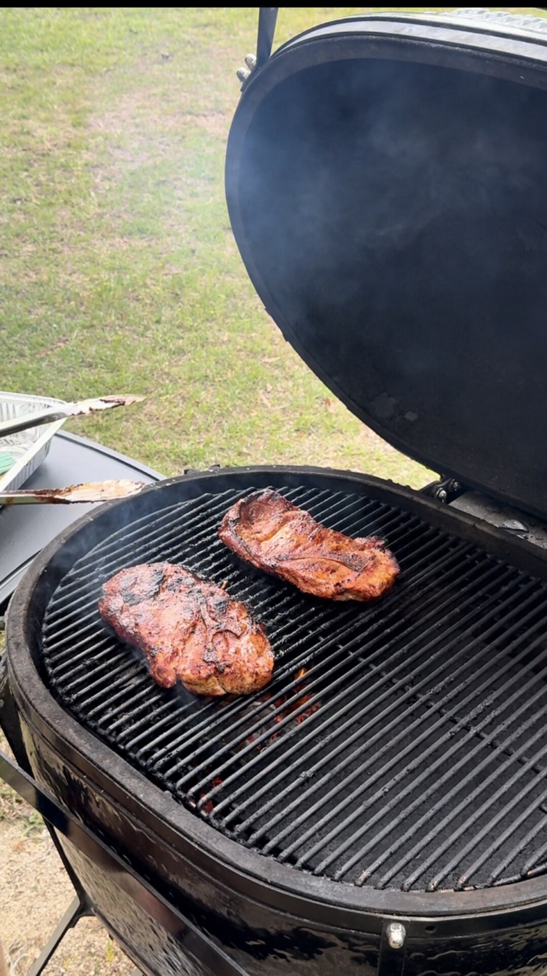 Two pieces of meat are grilling on a round charcoal barbecue with the lid open, while smoke rises and grass is visible in the background.