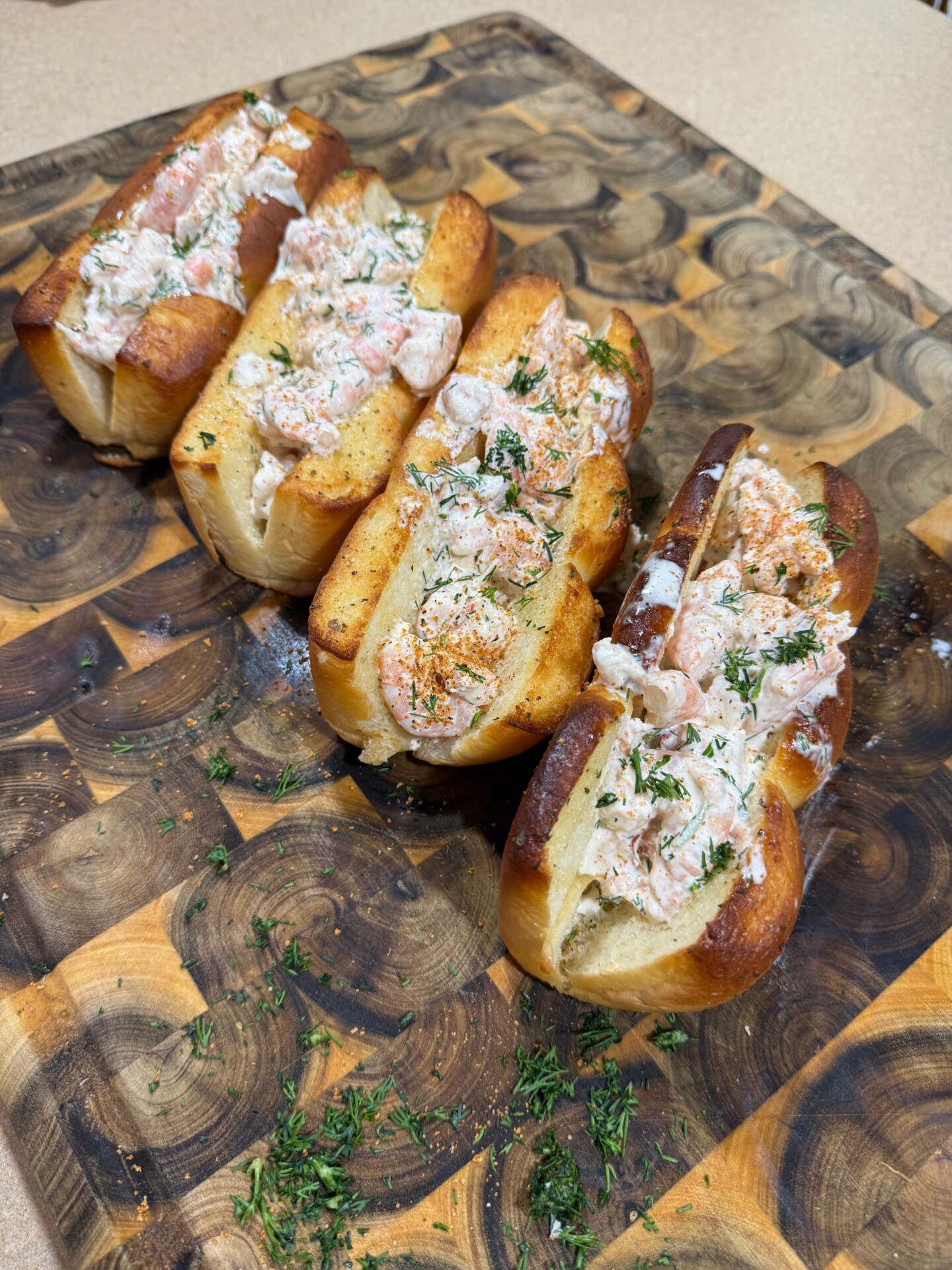 Three toasted buns filled with creamy lobster salad, topped with herbs and seasoning, are arranged on a wooden cutting board with scattered chopped herbs around them.