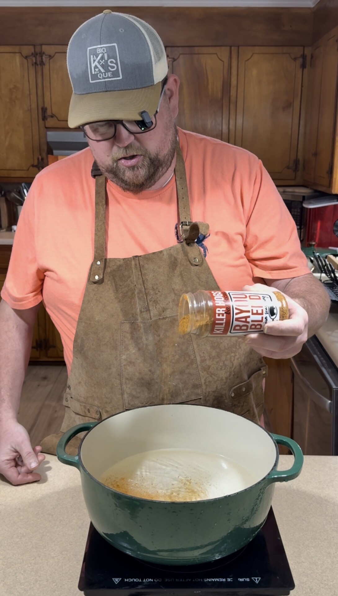A man wearing a hat, glasses, and an apron sprinkles Killer Hogs Bayou blend seasoning into a large green Dutch oven on a kitchen counter.