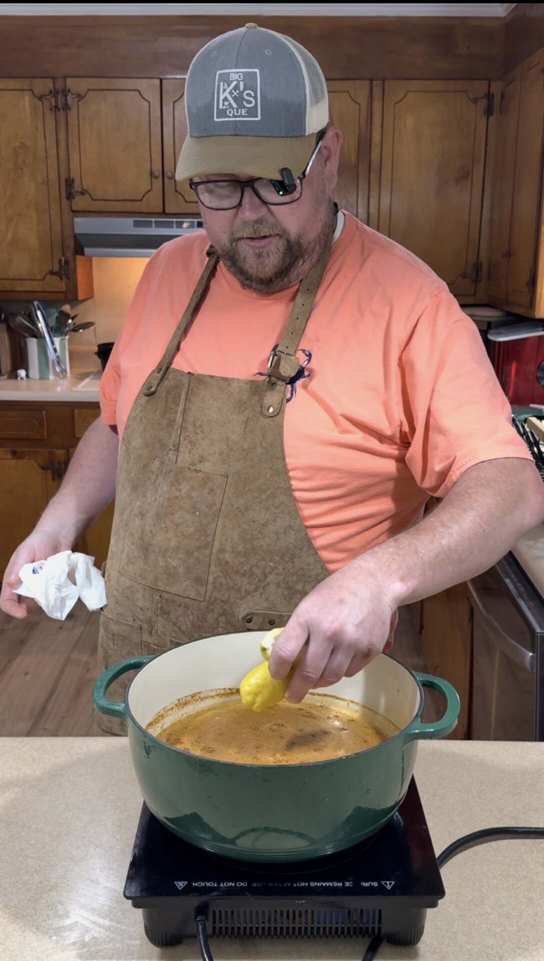 A man wearing an apron and cap stands in a kitchen, squeezing a lemon over a green pot on an electric stovetop, preparing a meal.