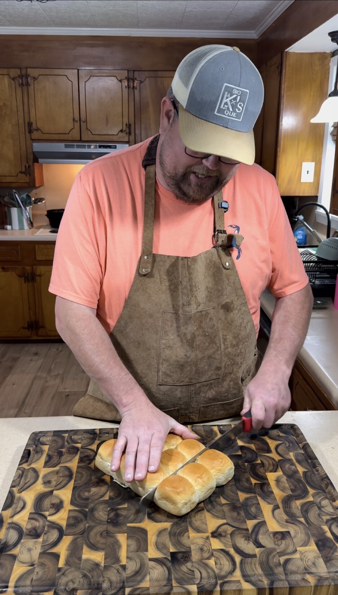 A man wearing a cap, glasses, an apron, and a coral shirt slices a cluster of bread rolls on a patterned wooden cutting board in a kitchen with wood cabinets.