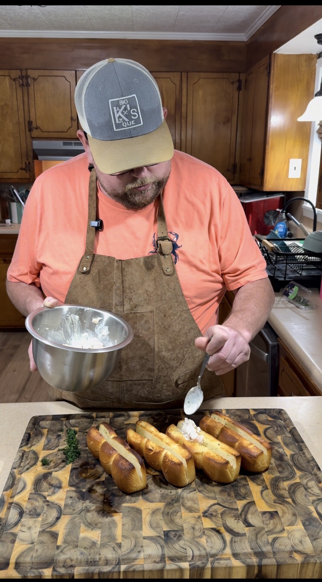 A man in a tan apron and cap is spooning a creamy mixture onto toasted sandwich rolls on a wooden cutting board in a kitchen with wood cabinets.