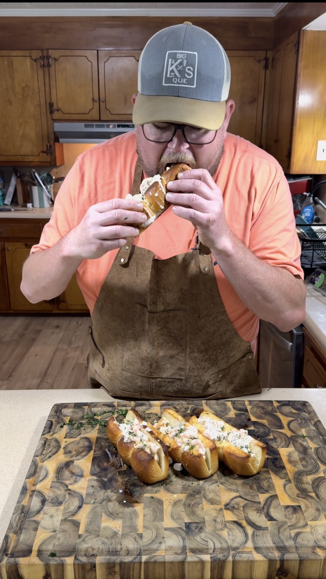 A man wearing a cap, glasses, and an apron is standing in a kitchen, eating a loaded hot dog. On the counter in front of him is a wooden board with more prepared hot dogs topped with cheese and herbs.