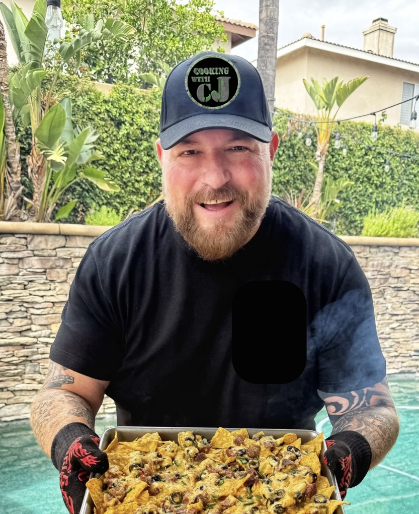 A bearded man in a black shirt and Cooking with CJ hat holds a steaming tray of nachos outdoors by a pool, smiling at the camera. Lush greenery and houses are visible in the background.