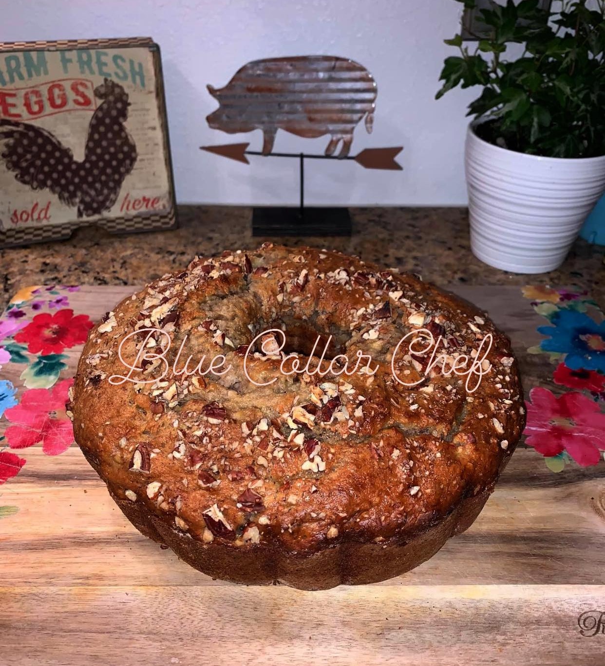 A homemade bundt cake topped with chopped nuts sits on a wooden board with a floral pattern. In the background, there’s a white potted plant, a metal pig decoration, and a vintage-style farm fresh eggs sign.