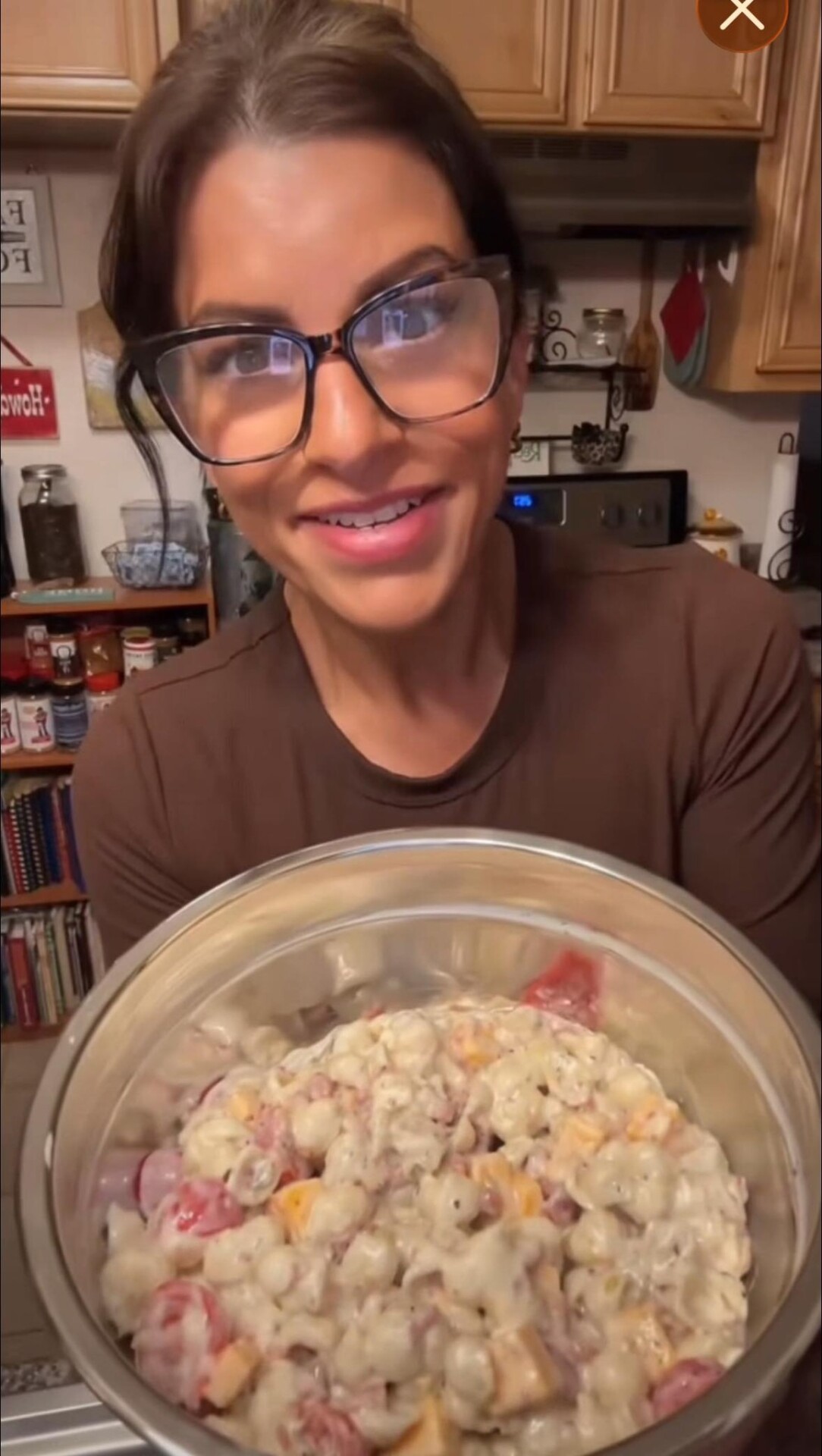 A woman with glasses and a brown shirt smiles while holding a large metal bowl filled with a creamy pasta dish with chunks of tomato and possibly cheese in a cozy kitchen setting.