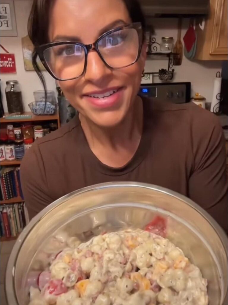 A woman wearing large glasses and a brown top is smiling and holding a bowl filled with creamy pasta salad with cheese and other ingredients in a cozy kitchen setting.