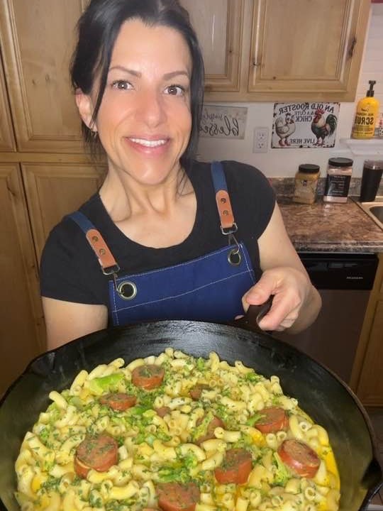 A woman in a blue apron smiles while holding a skillet filled with macaroni, sliced sausage, and garnished herbs in a kitchen with wooden cabinets.