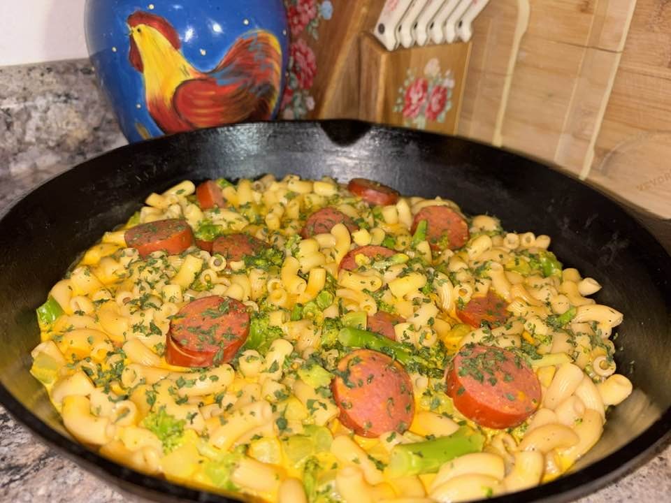 A skillet filled with macaroni pasta, sliced sausage, green bell peppers, and herbs sits on a kitchen counter near a ceramic rooster container and a wooden knife block.