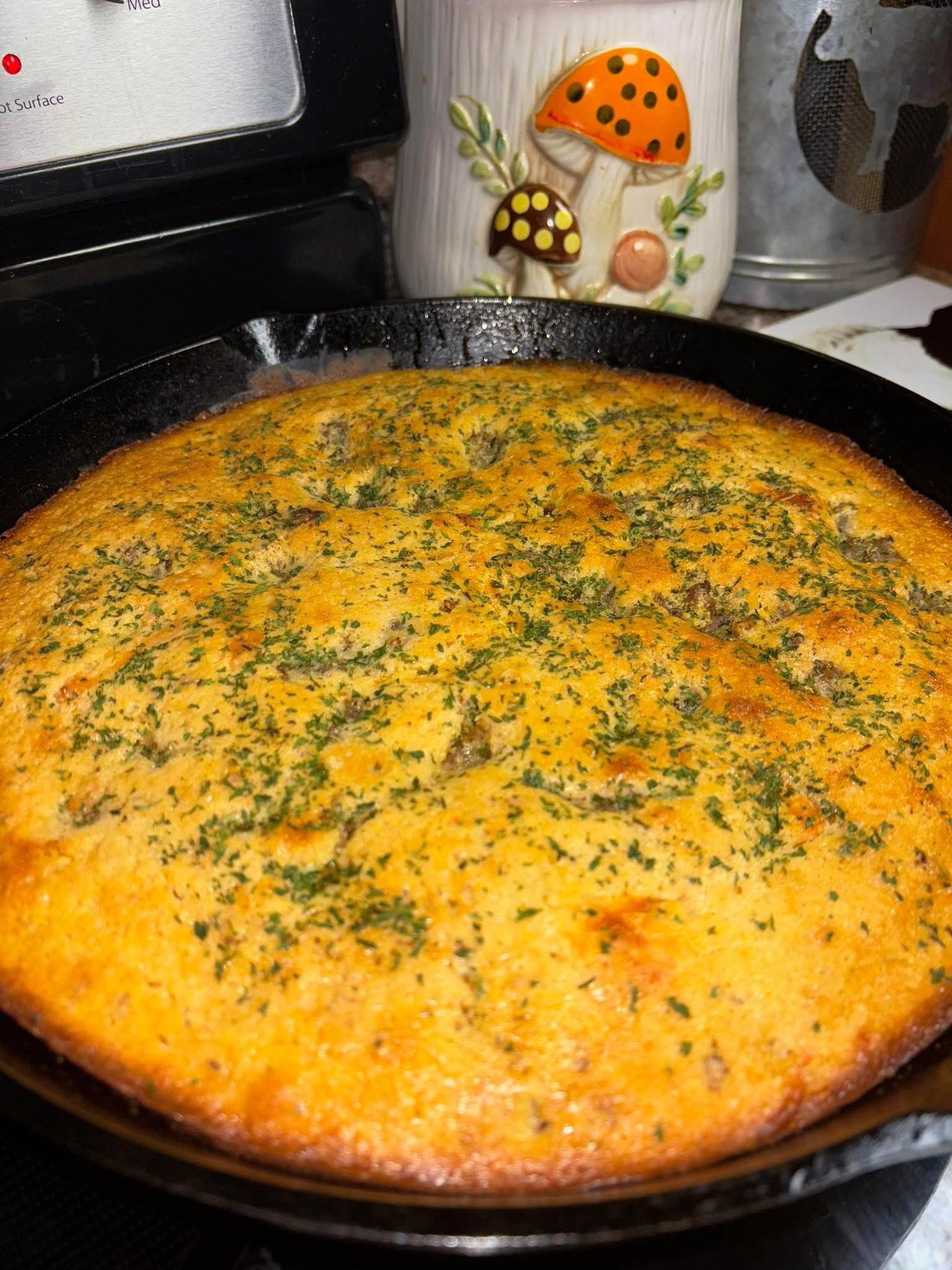 A golden-brown cornbread garnished with herbs in a black cast iron skillet sits on a stove. In the background, there is a decorative container with mushroom designs and a silver kitchen canister.