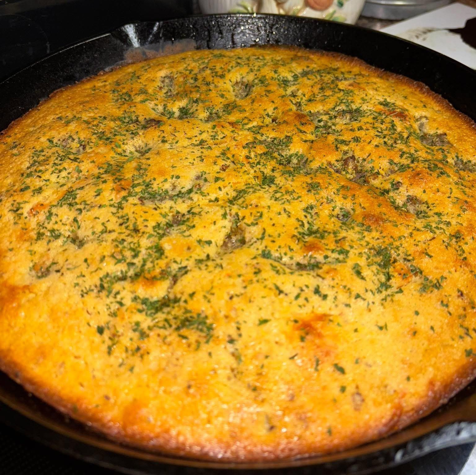 A freshly baked, golden-brown cornbread topped with herbs is shown in a cast-iron skillet on a stovetop. The surface looks slightly crisp with visible green flecks of seasoning.