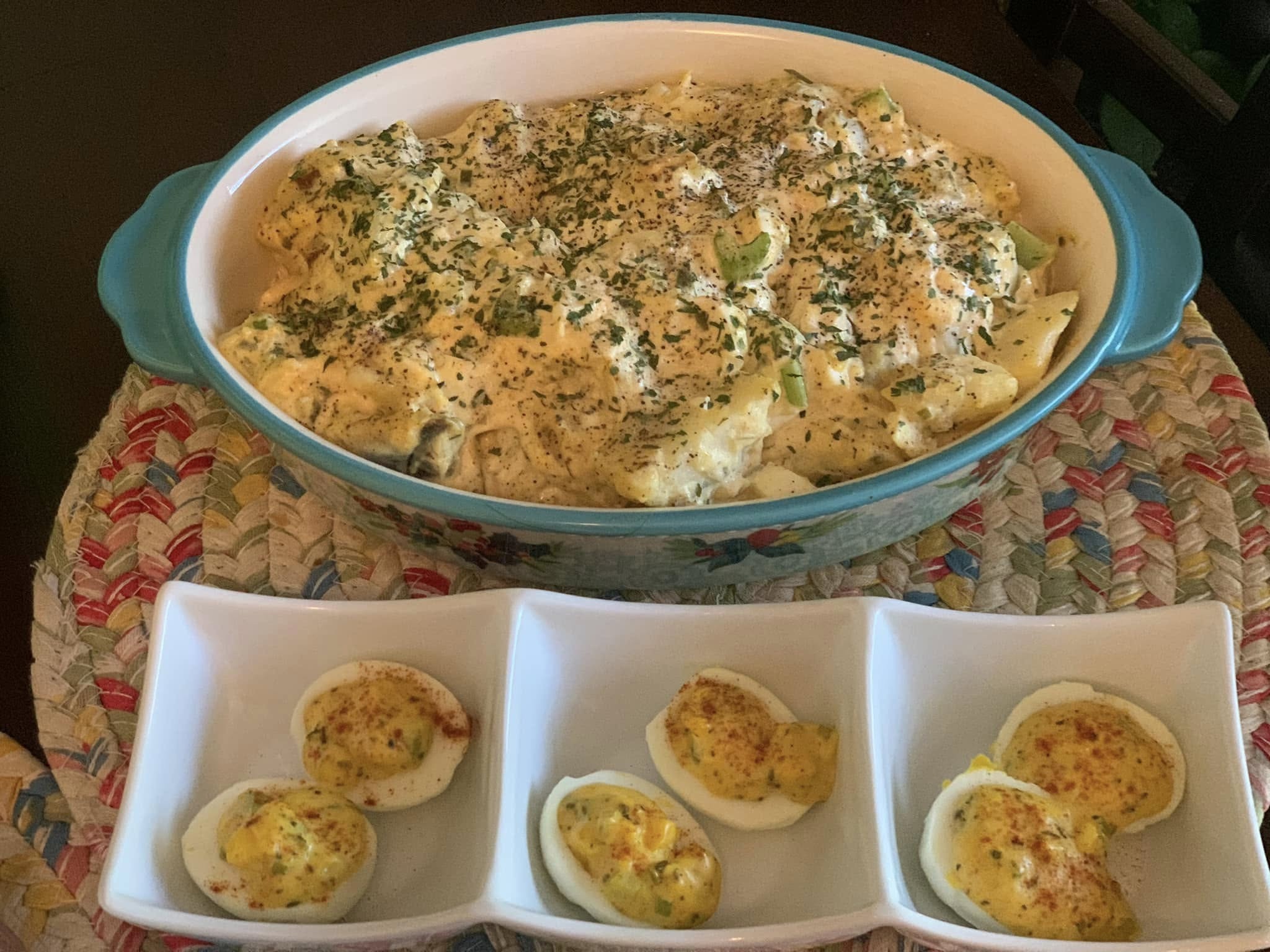 A blue-rimmed baking dish filled with creamy pasta topped with herbs, placed on a woven mat. In front, three deviled eggs are arranged in a white divided dish, garnished with spices.
